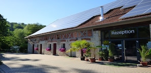 A building with a reception area, solar panels on the roof, and potted plants around the entrance. - Camping Schwabenmühle (Weikersheim)