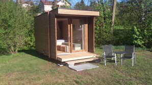A wooden cabin with a sliding glass door, a small porch, and two metal chairs. - Camping Schwabenmühle (Weikersheim)