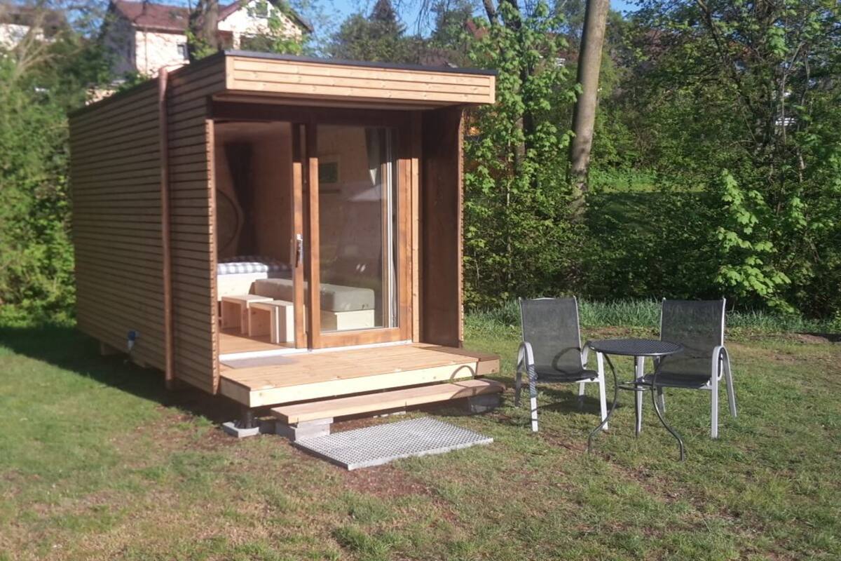 Une cabane en bois avec une porte coulissante en verre, un petit porche et deux chaises en métal.