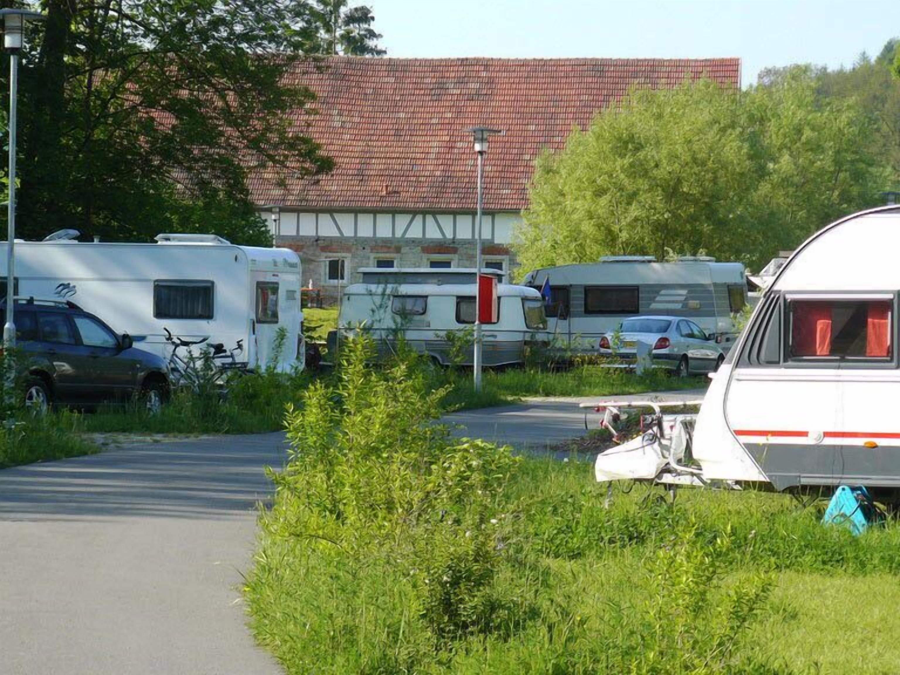 En campingplads med autocampere parkeret på en belagt overflade, omgivet af grønt og en bygning med røde tegltag i baggrunden.