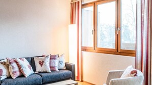 A cozy living room with a sofa and armchair, a window with curtains, and a radiator. - Titlis Resort (Engelberg)