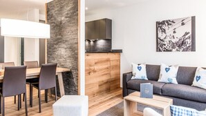A modern living room with a sofa, dining table, and chairs. There is a kitchen area with cabinets and a stove. A painting of a mountain landscape is on the wall. - Titlis Resort (Engelberg)