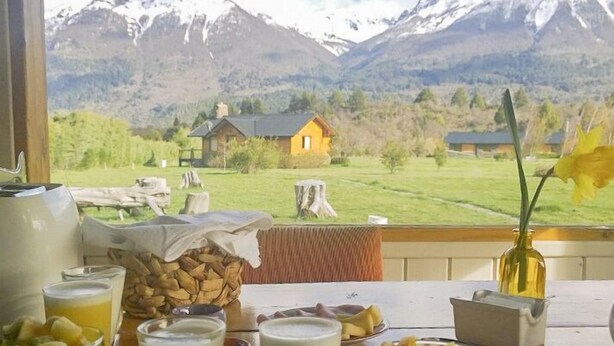 Une table de petit-déjeuner avec divers aliments et boissons, un vase rempli de fleurs, et une vue sur des montagnes et une maison.