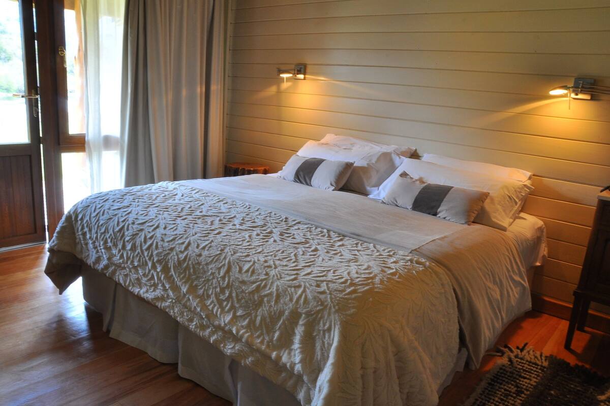 Una cama bien hecha con colcha y almohadas estampadas, suelo de madera y una lámpara fijada a la pared.