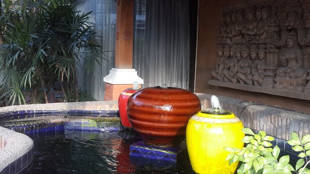 Una fontana decorativa con un vaso rosso e giallo di grandi dimensioni, circondata da una piscina in mosaico e piante in vaso.