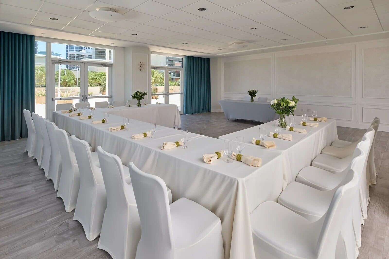 A conference room with a long table set for a meeting, white chairs, and a view of the outdoors through large windows.