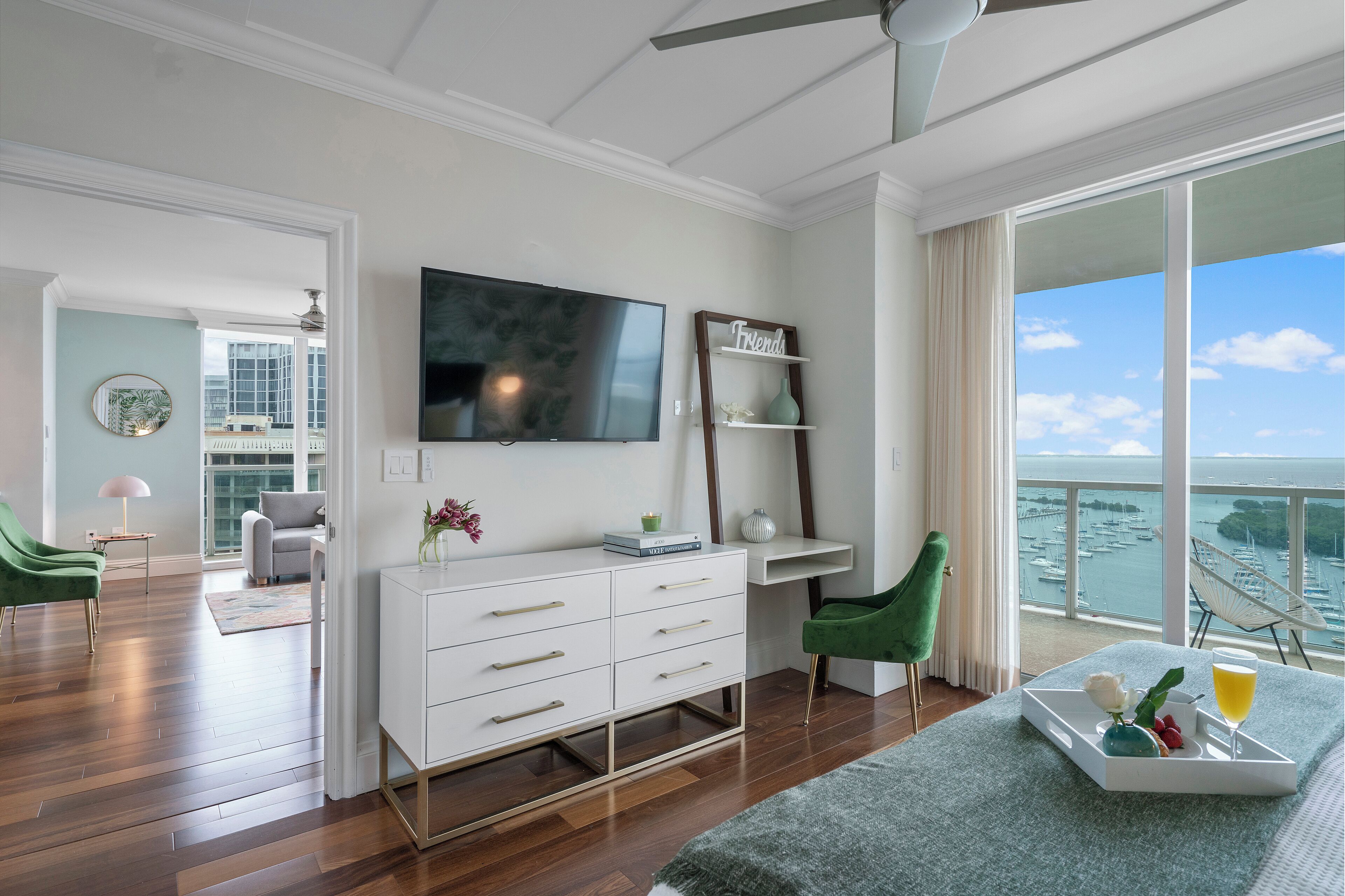 A modern hotel room with a large balcony, a flat-screen TV, a desk, and a dining area with a tray of fruit and a drink.