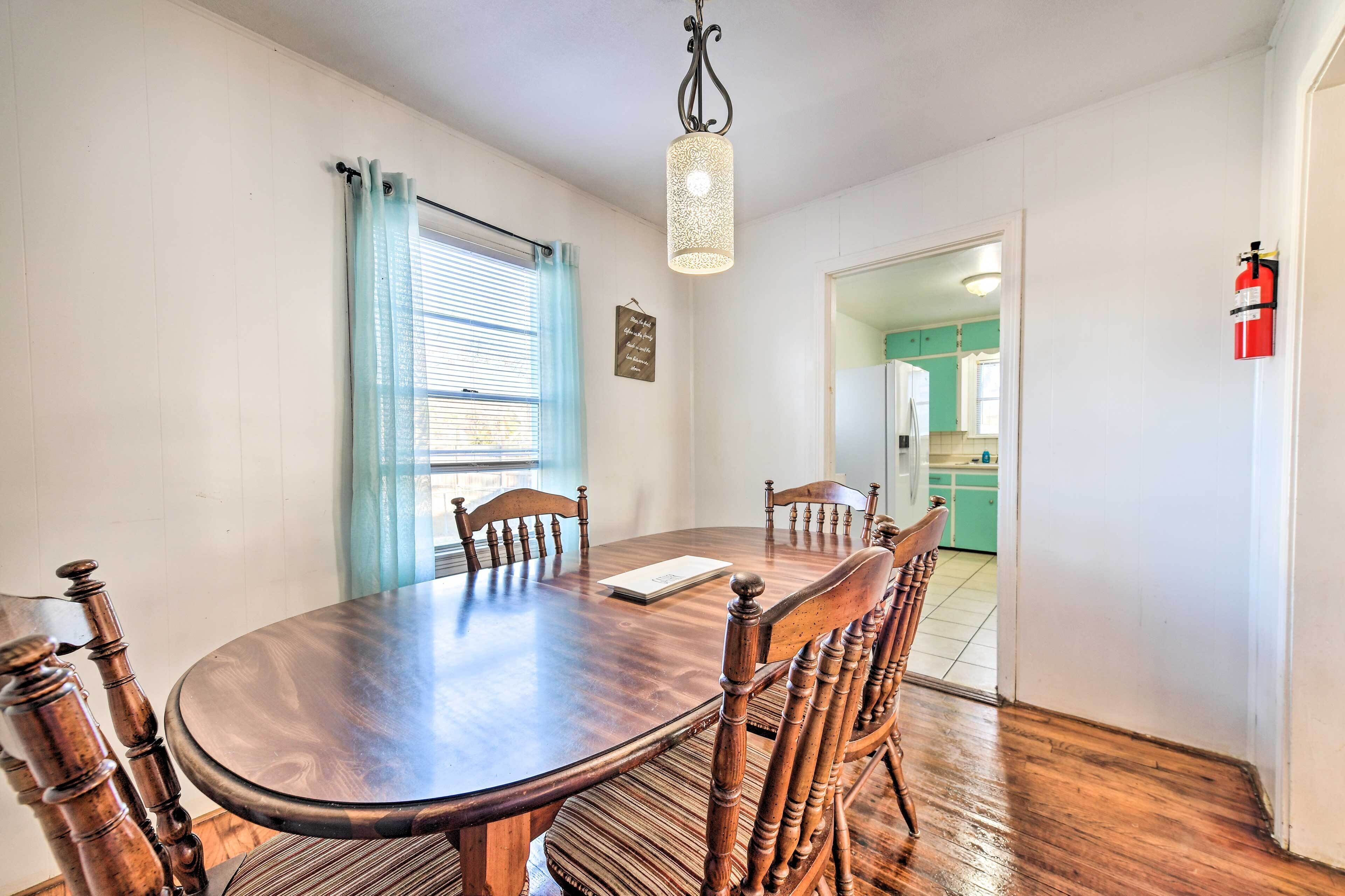 A dining area with a wooden table and chairs, a hanging light fixture, a window with blue curtains, and a kitchen visible through an open door.