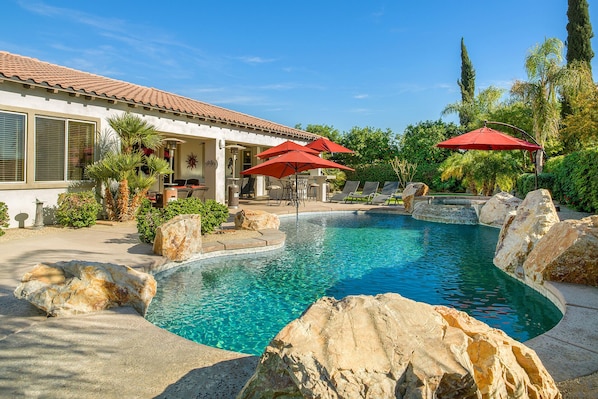 Une piscine au design incurvé, entourée de rochers et de palmiers. Des espaces de détente extérieurs sont aménagés avec des parasols rouges.