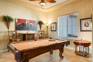 Une table de billard avec une queue de billard, des chaises et une lampe dans une pièce dotée d’un ventilateur de plafond, de tableaux encadrés et d’une fenêtre avec des stores.