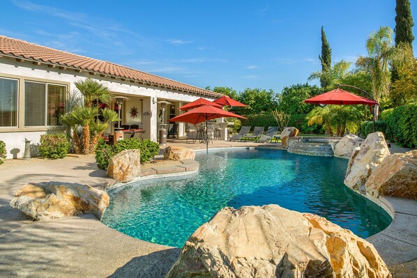 A swimming pool with a curved design, surrounded by rocks and palm trees. There are outdoor seating areas with red umbrellas.