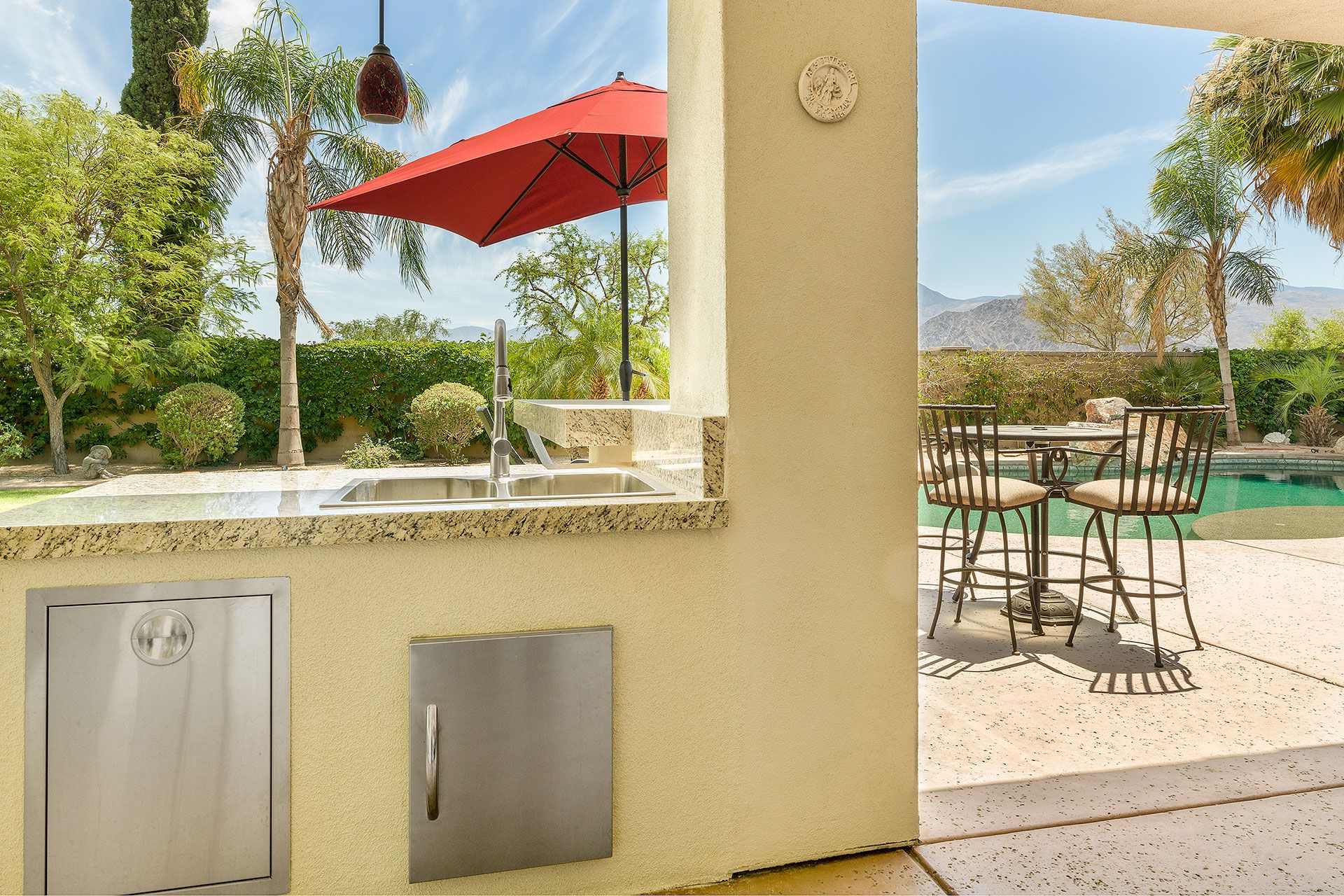 An outdoor kitchen area with a sink, a red umbrella, and a poolside seating area.
