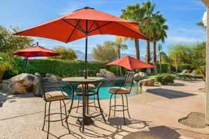 A poolside area with a round table, two bar stools, and two red umbrellas. - Villa Blanca - Desert Getaway w/ 6 Bedrooms! (Indio)