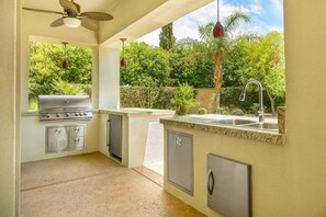 An outdoor kitchen with a grill, sink, and storage cabinets, surrounded by greenery.