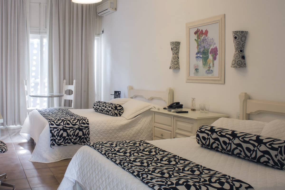 A hotel room with two beds, a desk, a chair, a window with curtains, an air conditioning unit, and a framed floral artwork on the wall.
