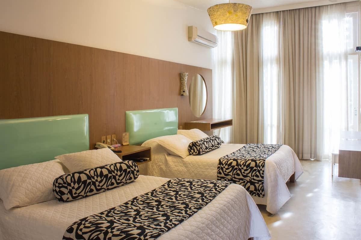 A hotel room with two beds, a bedside table with a lamp, a mirror, and a window with curtains.