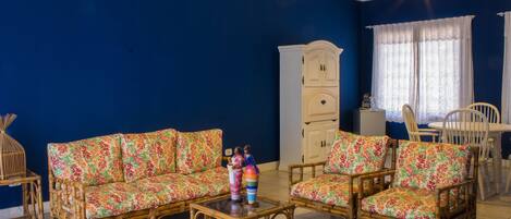 Una sala de estar con pared azul, un mueble blanco, un sofá de mimbre, una mesa de centro de madera y una mesa de comedor con sillas.