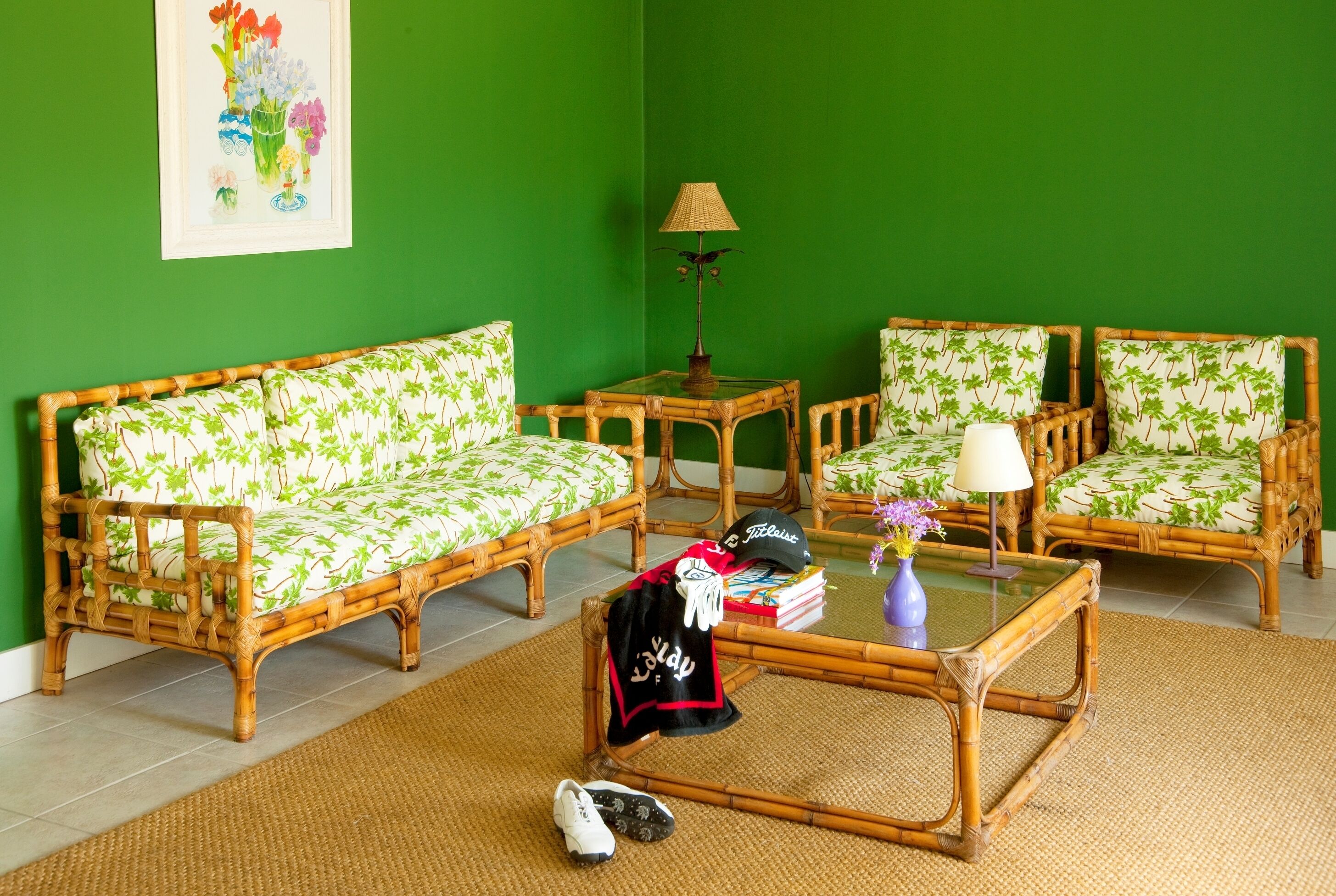 A room with a green wall, a rattan sofa, two rattan chairs, a glass coffee table, and a floor lamp.
