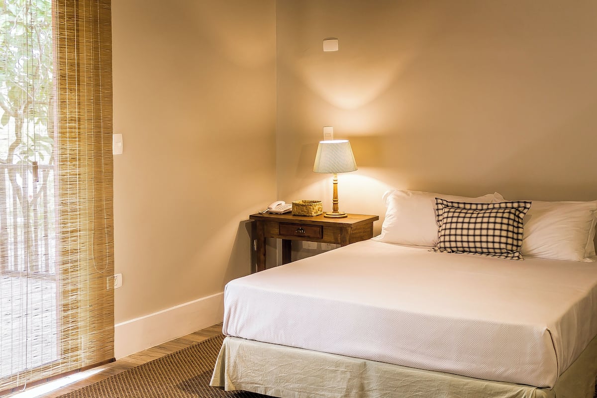 A neatly made bed with a checkered pillow, a bedside lamp, and a small wooden nightstand with a lamp and a phone.