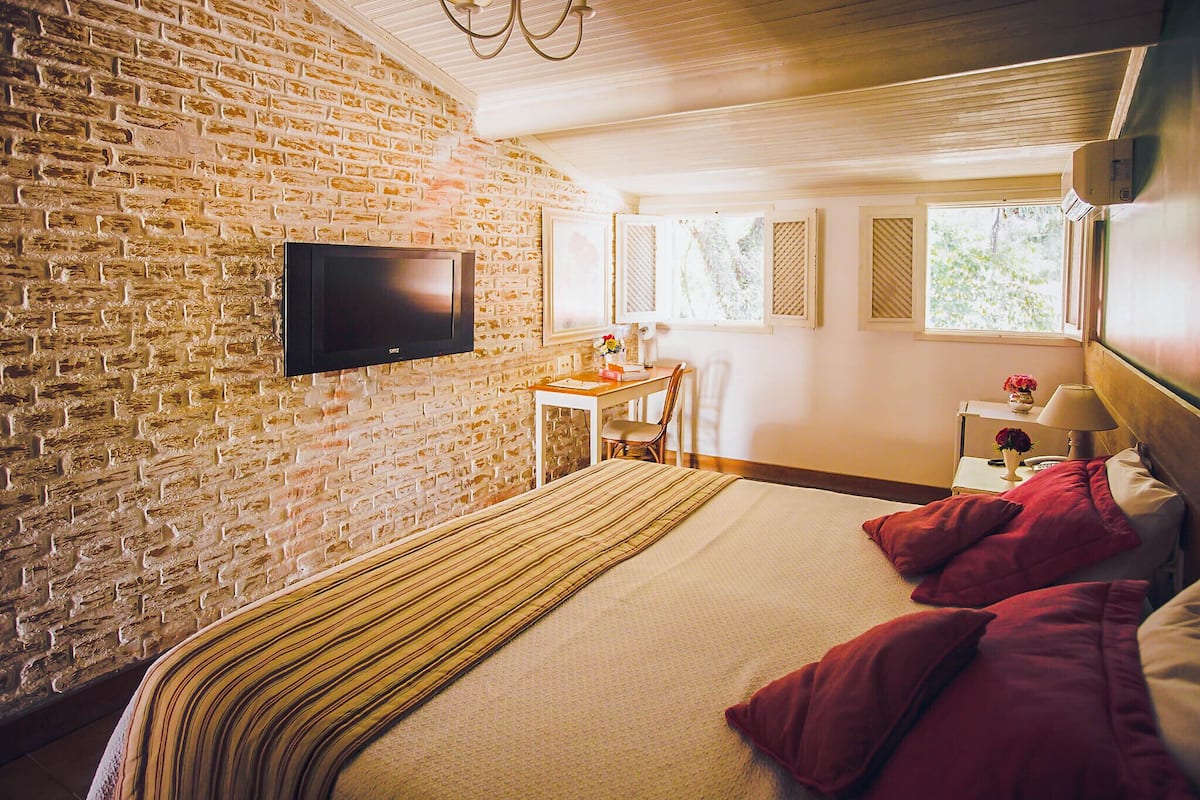A room with a brick wall, a flat-screen TV mounted on it, a bed with red pillows, a wooden nightstand with a lamp, and a window with shutters.