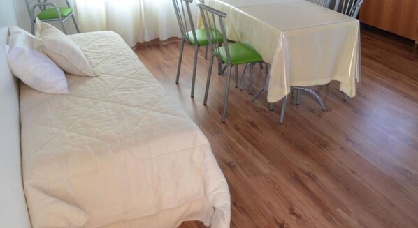 A single bed with a quilt, a table with a tablecloth, and two green chairs.