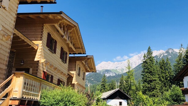 Un chalet en bois doté d’un balcon, situé dans un cadre magnifique, avec des montagnes enneigées en toile de fond et une végétation luxuriante.