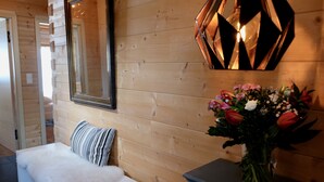Un mur en bois avec un miroir, un luminaire aux formes géométriques, un banc recouvert d’un plaid en fausse fourrure blanche et un vase rempli de fleurs.