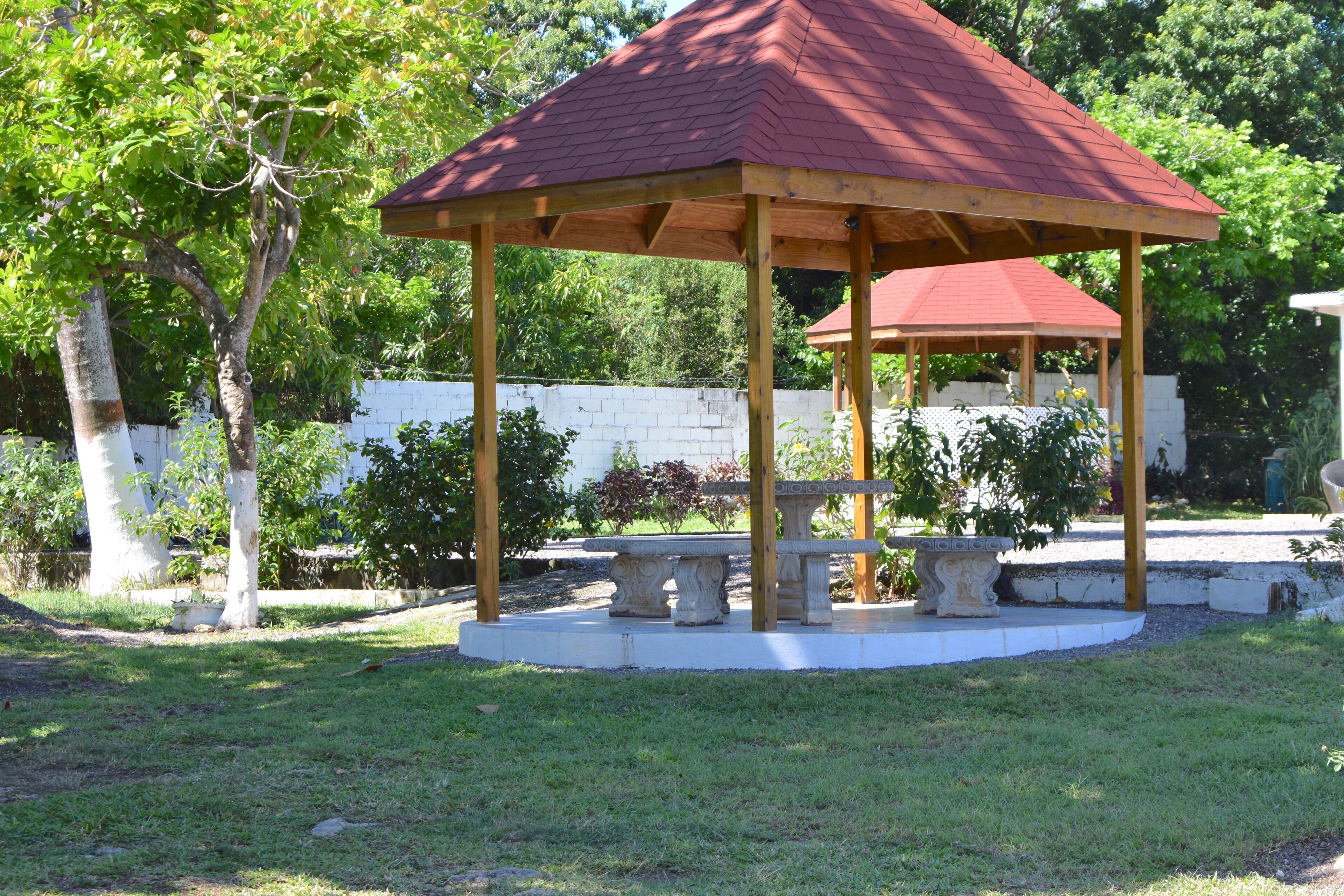 A gazebo with a red roof and wooden beams, surrounded by a grassy area and trees.