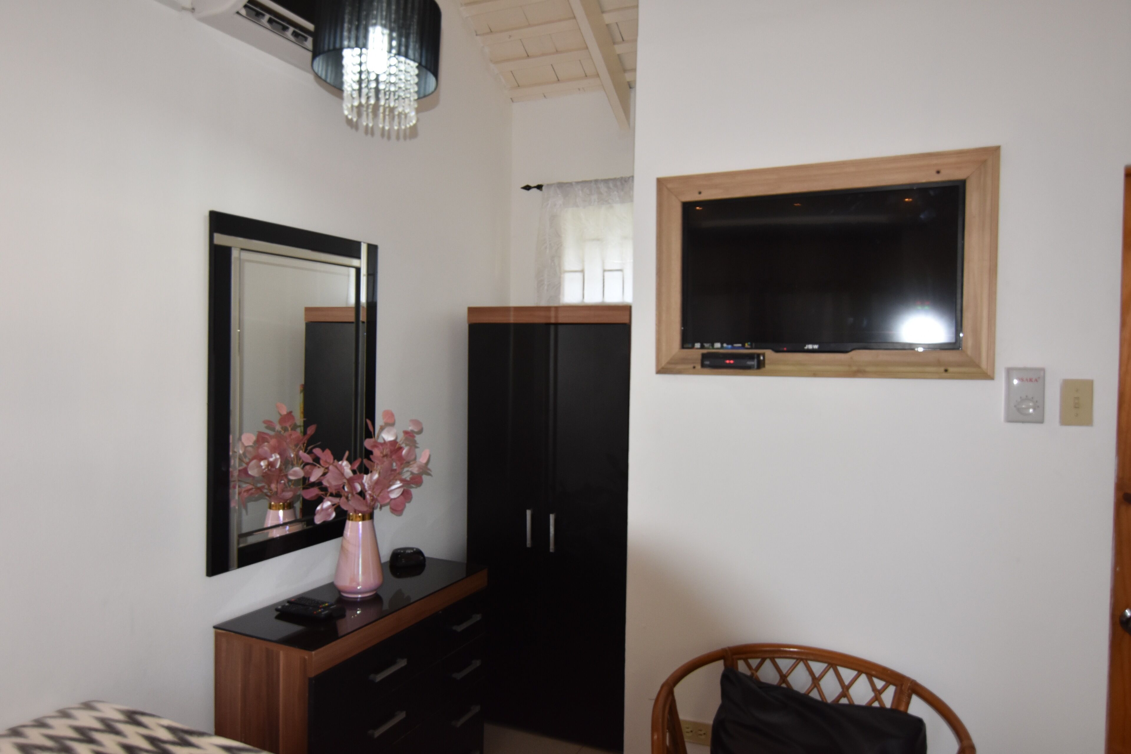 A room with a large mirror, a wooden cabinet, a black wardrobe, a small table with a vase, a wooden chair, and a window with a wooden frame.