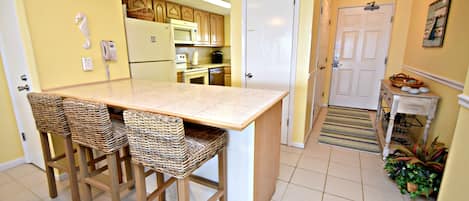A kitchen with a central island, bar stools, a microwave, a refrigerator, and a door leading to another room.