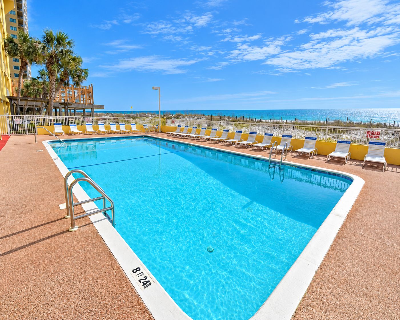 Une piscine rectangulaire à bordure blanche, entourée de chaises longues et d’une rambarde.