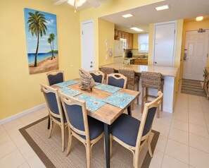 Un coin repas avec une table et des chaises en bois, une cuisine à l’arrière-plan et un tableau représentant une scène de plage accroché au mur.