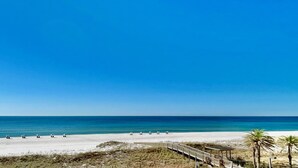 Una playa con un paseo de madera que conduce a un área techada, palmeras y un cielo azul despejado.