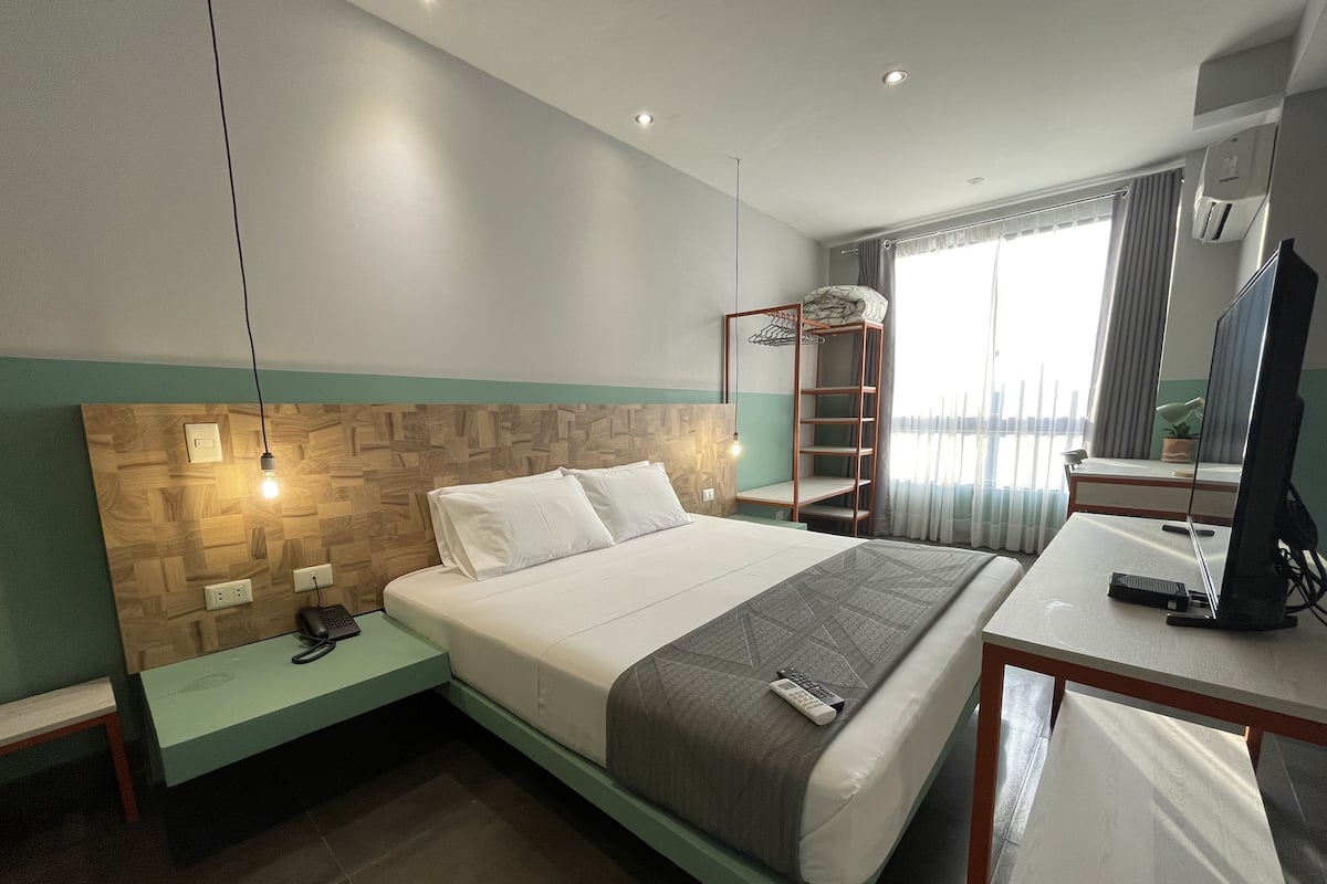 A modern hotel room with a neatly made bed, a desk with a computer, a small bedside table with a lamp, and a window with curtains.