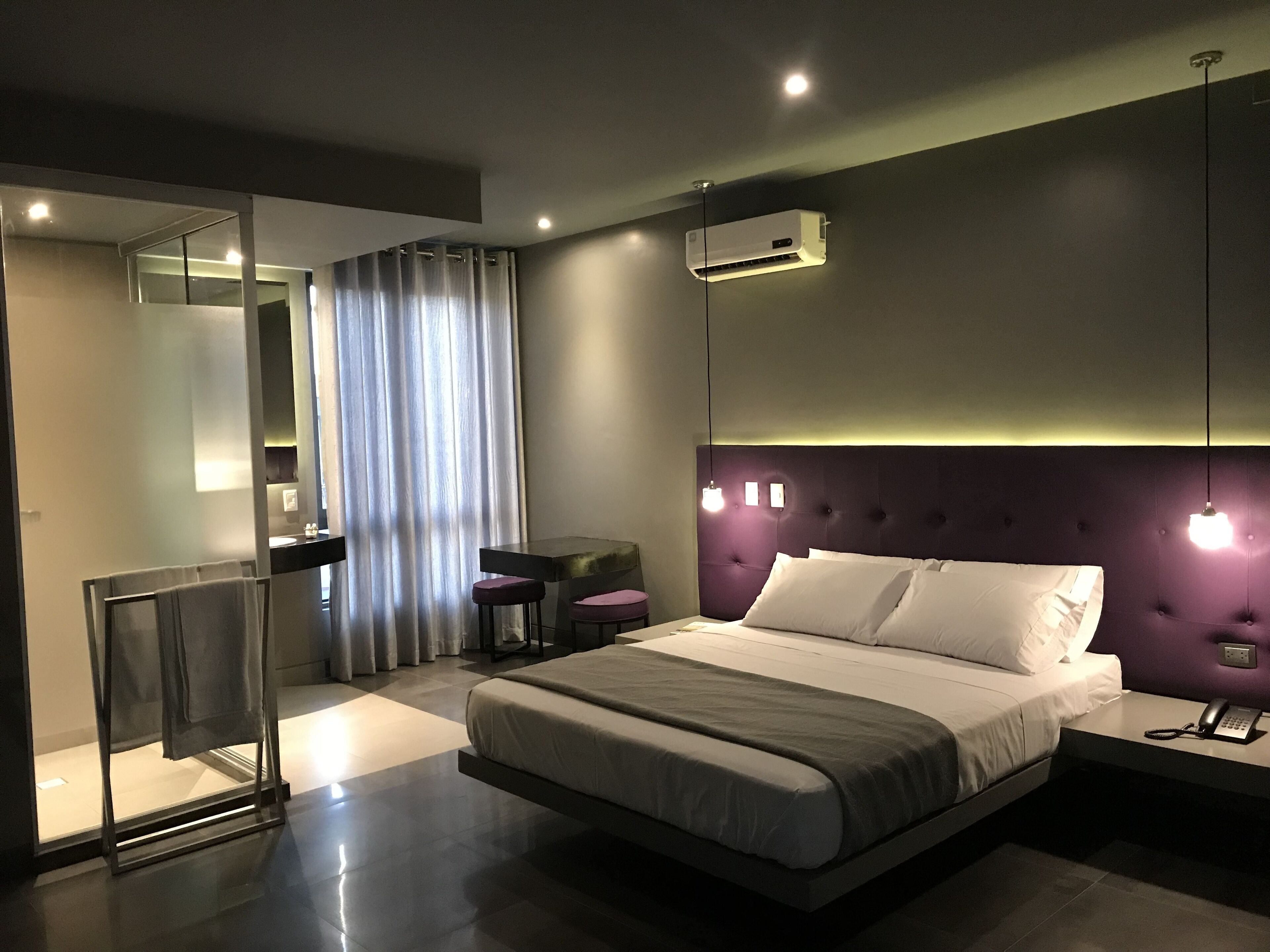 A modern hotel room with a large bed, a bedside table with a lamp, a mirror, a window with curtains, and an air conditioning unit.