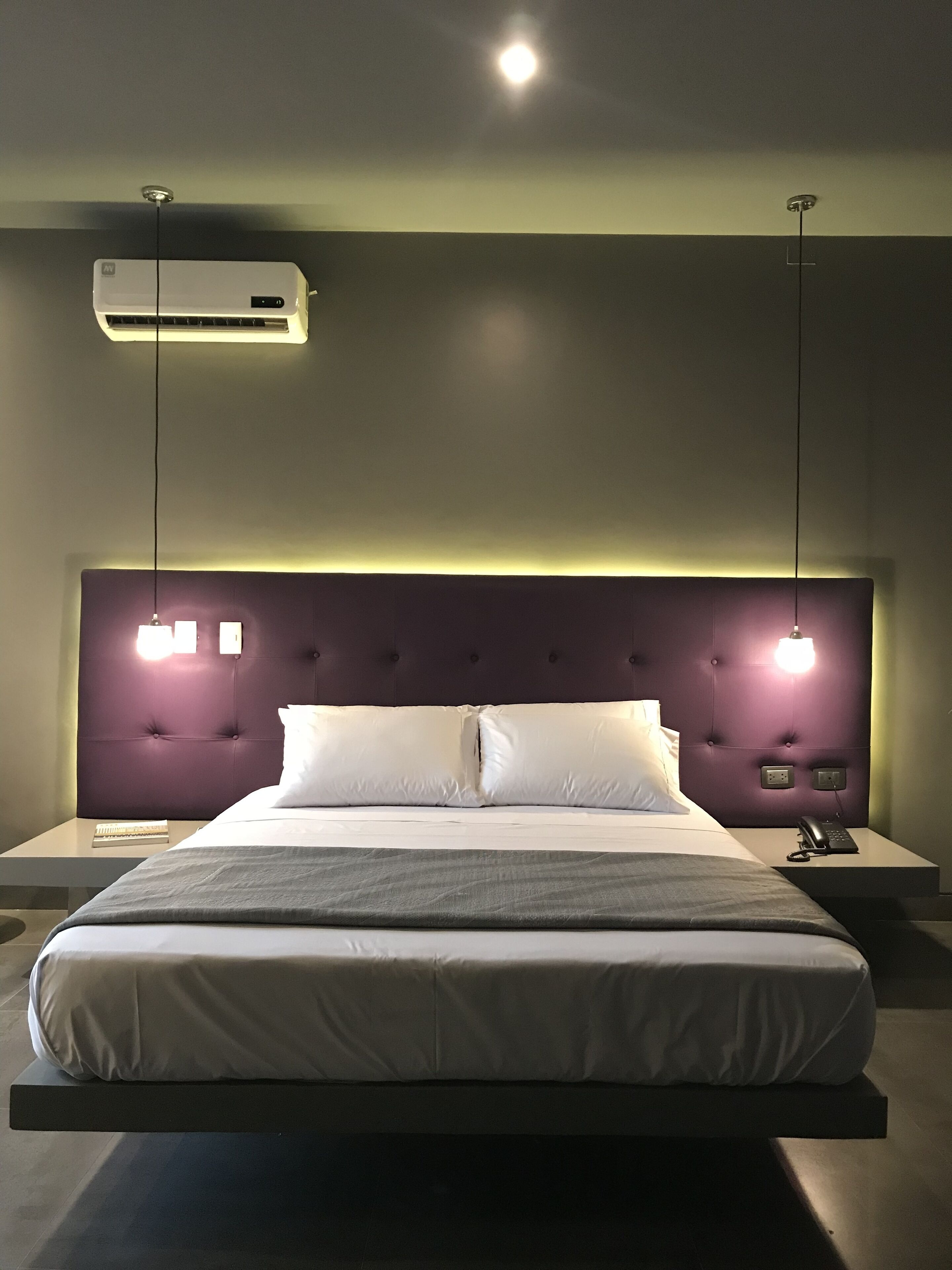 A modern hotel room with a large bed, a headboard with integrated lighting, a bedside table with a phone, and an air conditioning unit.