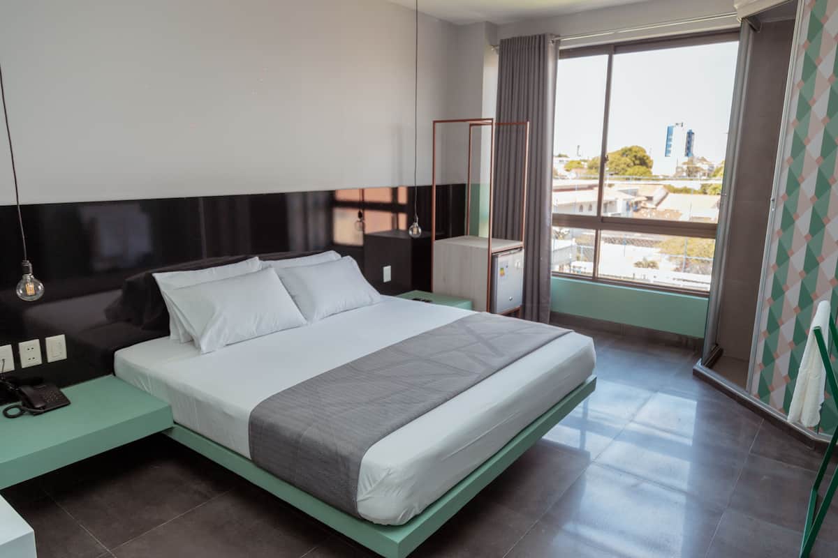 A modern hotel room with a large bed, a bedside table with a lamp, a nightstand, and a view of the city through a large window.