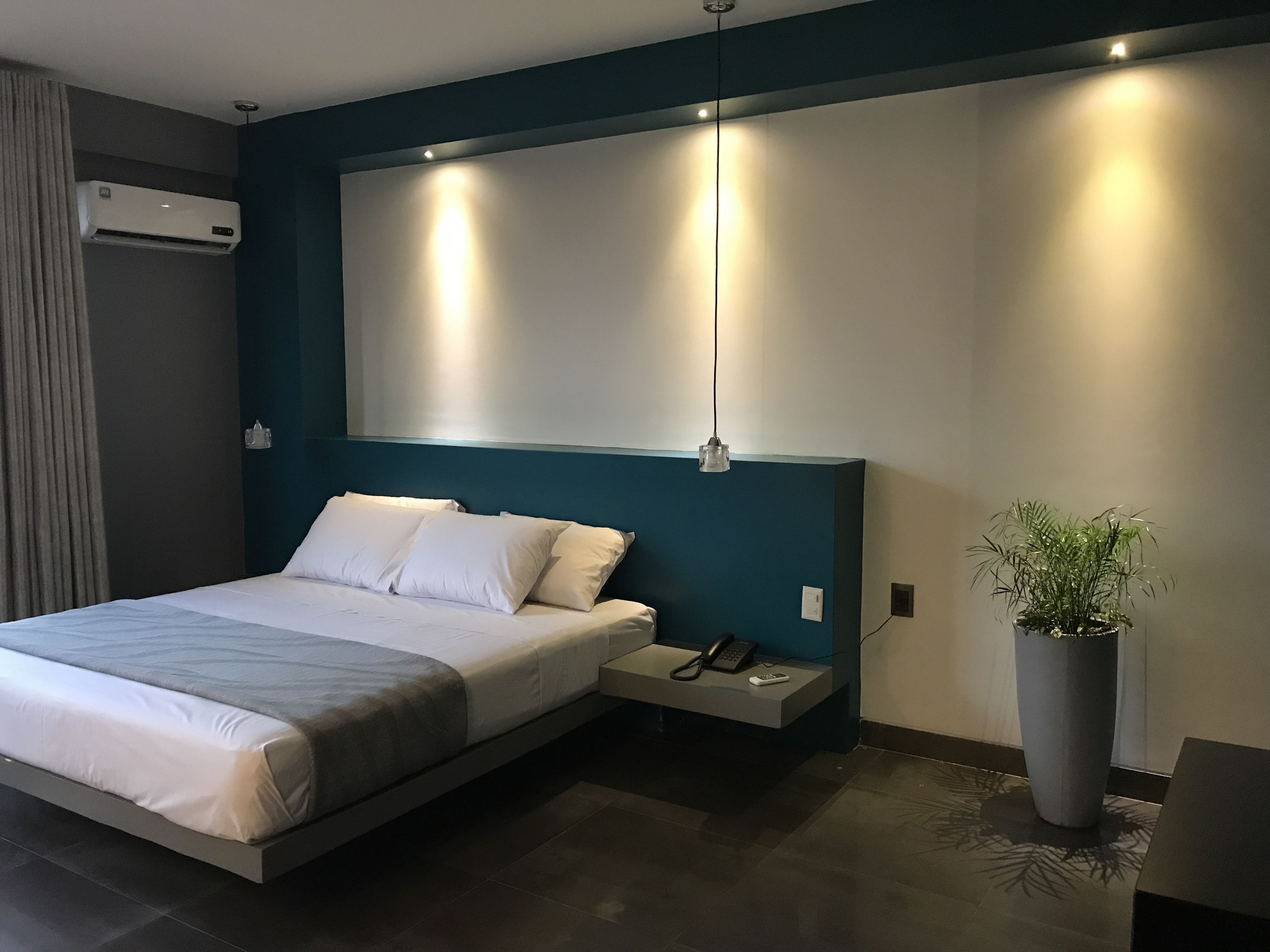 A modern hotel room with a bed, bedside table with a phone and remote, a potted plant, and a wall-mounted air conditioner.