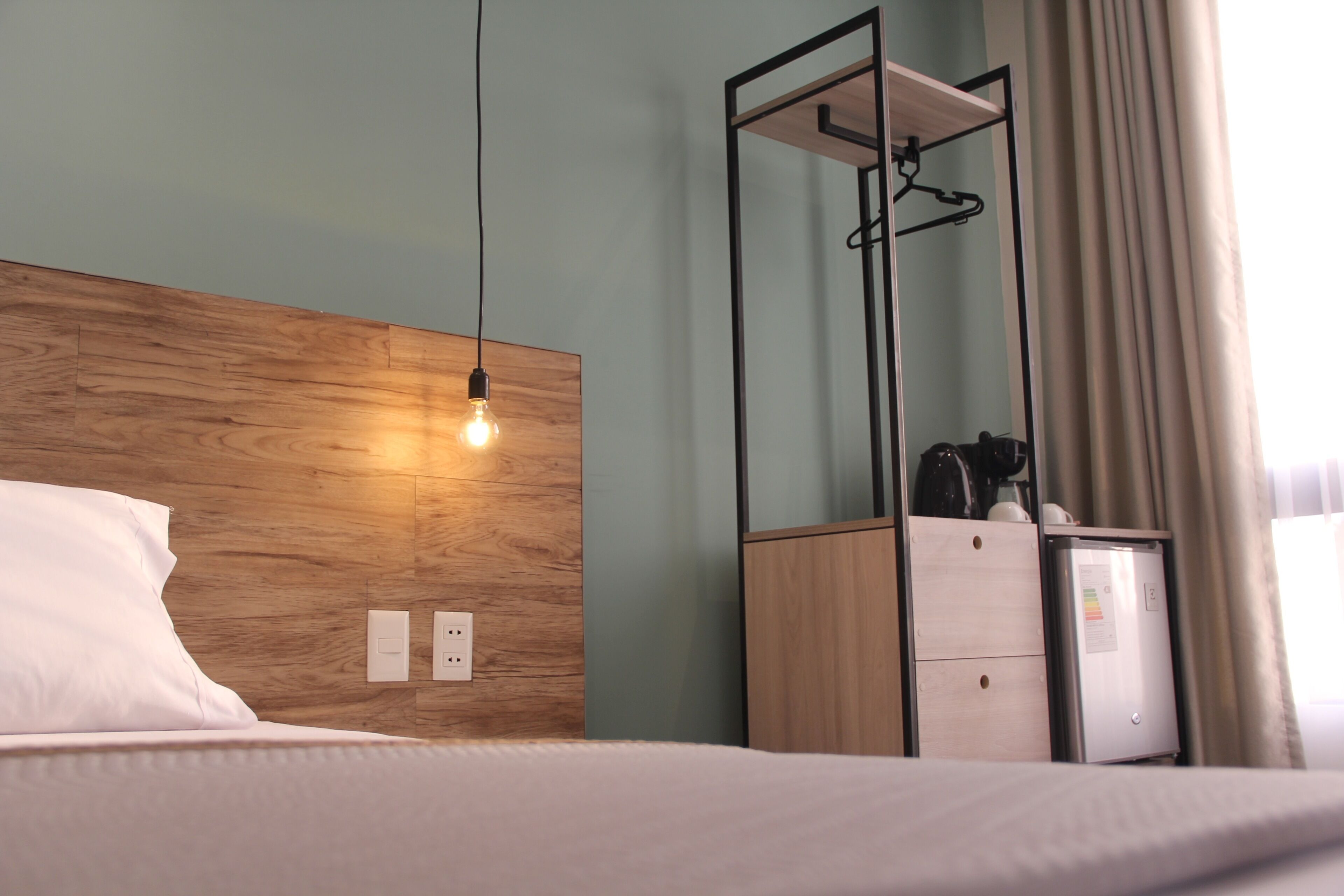Ein Hotelzimmer mit einem hölzernen Kopfteil, einem Bett mit weißer Bettwäsche, einem kleinen Schreibtisch mit Kaffeemaschine und einem Spiegel mit Kleiderhaken.