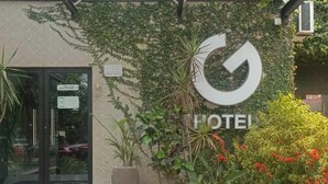 Een hotelentree met een glazen deur, een groot logo en een verzorgde tuin.