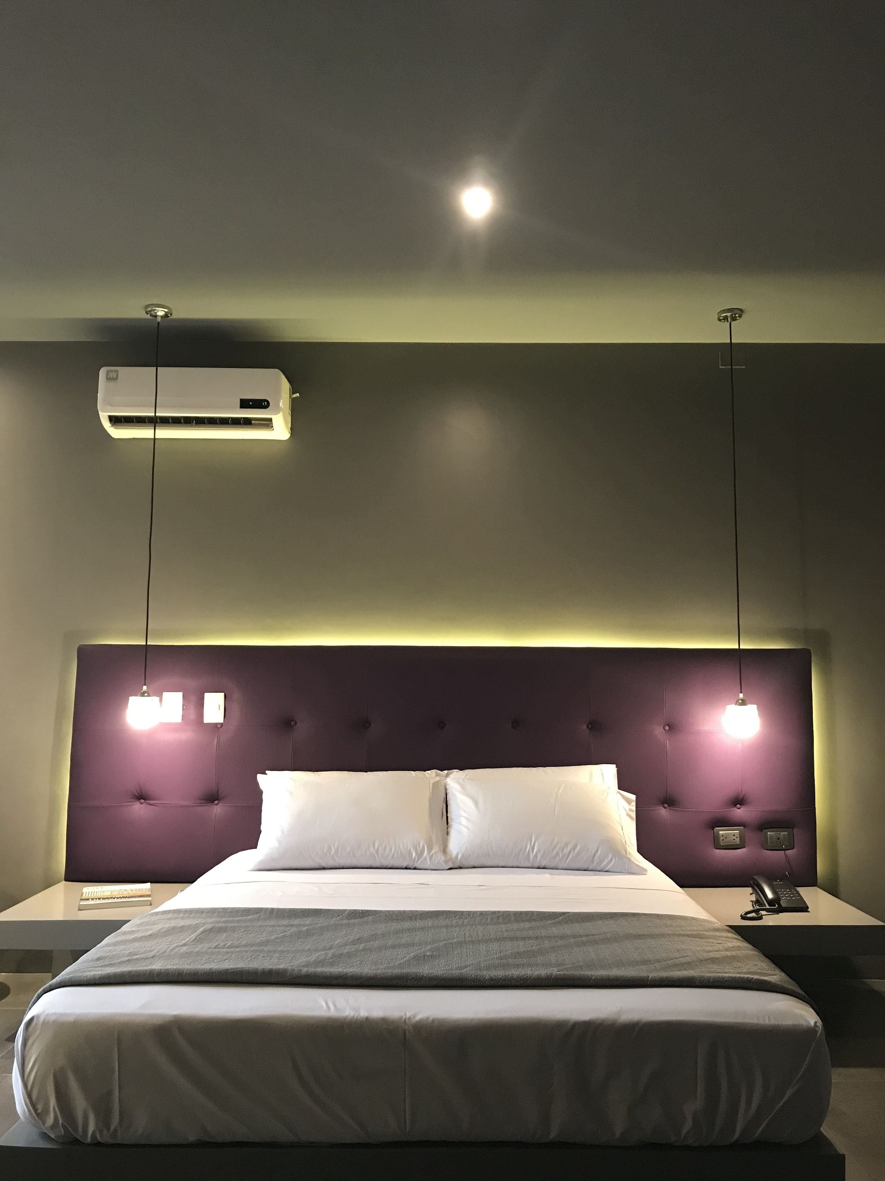 A modern hotel room with a large bed, a headboard with integrated lighting, an air conditioning unit, and a bedside table with a phone and a lamp.