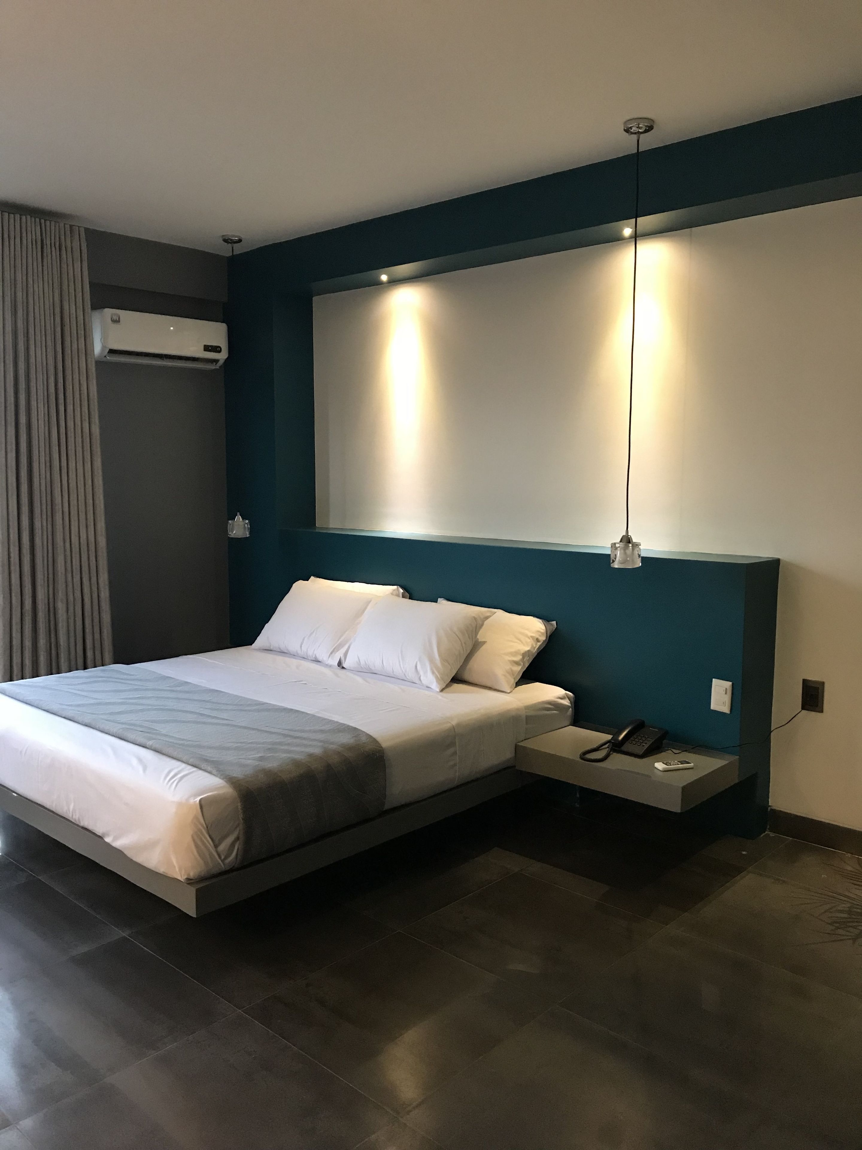 A modern hotel room with a large bed, bedside table with a phone and remote, and an air conditioning unit on the wall.