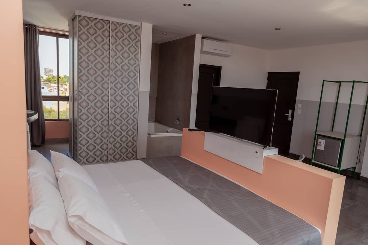 A modern hotel room with a bed, a TV, a small bathroom, and a view of the city.