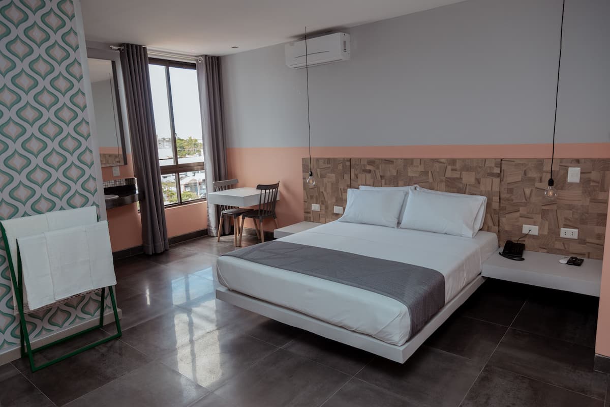 A modern hotel room with a large bed, a small desk, a chair, and a window with curtains.