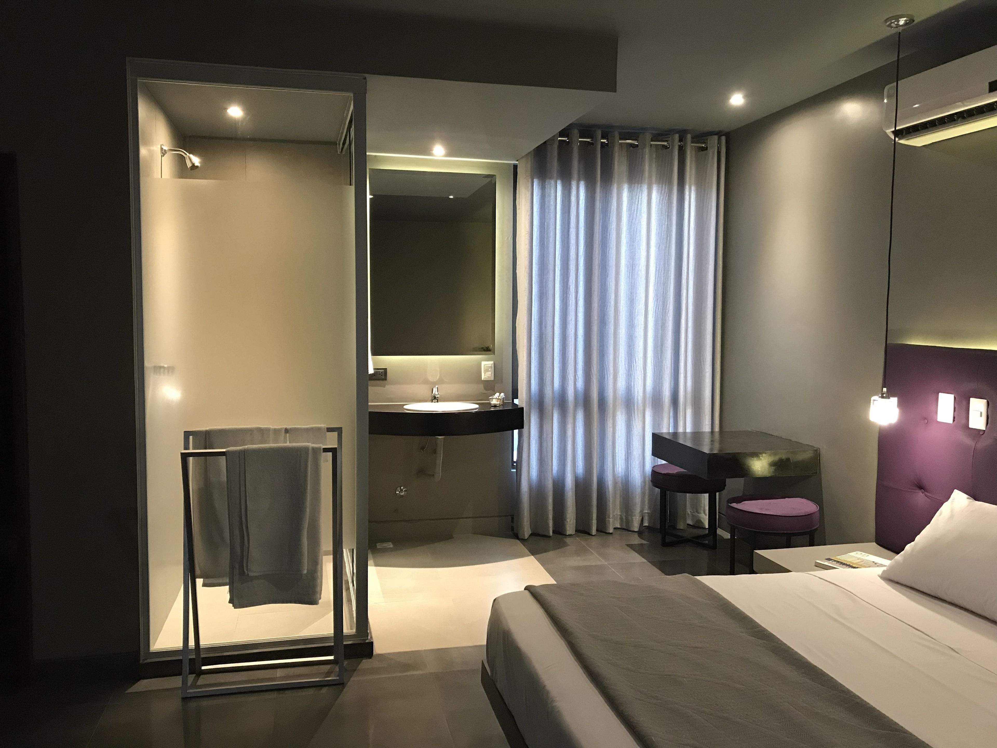 A modern hotel room with a walk-in shower, a vanity with a mirror, a bed with a headboard, and a desk with a chair.