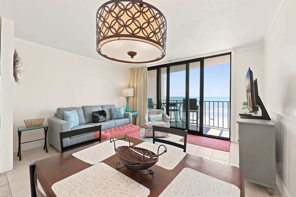 A room with a dining table, a sofa, a chair, a lamp, a TV, and a view of the sea. - Oceanfront in Heart of N Myrtle  Beach with Pool (North Myrtle Beach)