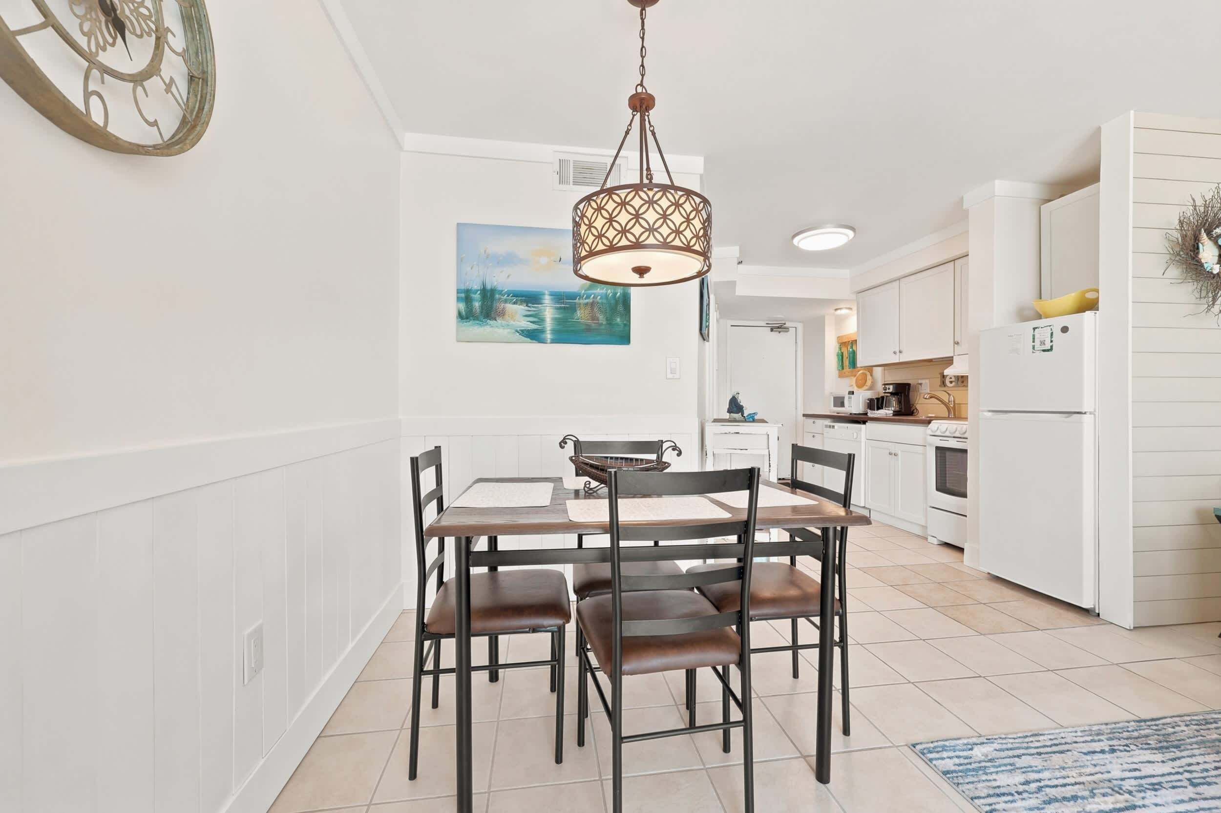 A dining area with a table and chairs, a hanging light fixture, a painting on the wall, and a kitchen in the background.