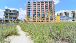 Un hotel sulla spiaggia con diversi piani, un viale sabbioso e vegetazione circostante.