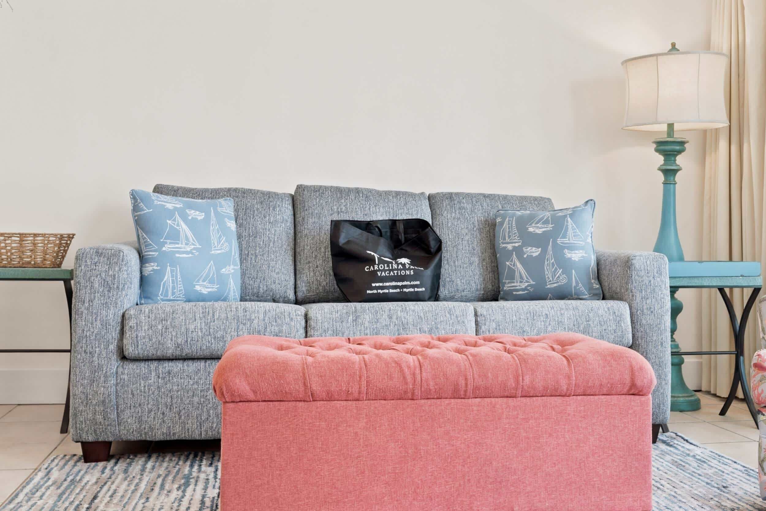 A grey sofa with patterned pillows, a teal lamp, and a pink ottoman in a room with a woven basket and a small table.