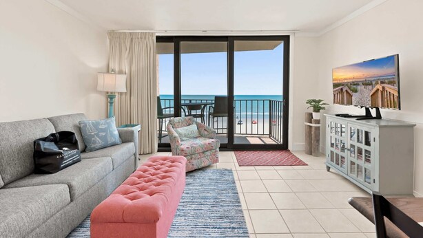 Un soggiorno con divano, poggiapiedi rosa, una poltrona, televisore, mobile contenitore e un balcone vista mare.